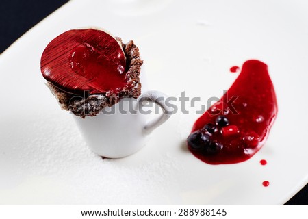Chocolate cake baked in a mug decorated with chocolate circular plate, painted in red stripes, berry jam, black and red currants on a white plate on a black background