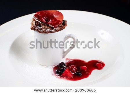 Chocolate cake baked in a mug decorated with chocolate circular plate, painted in red stripes, berry jam, black and red currants sprinkled with powdered sugar on a white plate on a black background