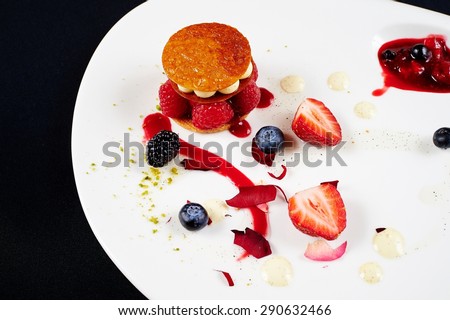 Original unusual berry dessert with honey tuille, raspberry and cream, decorated with strawberries, blueberries, cream, BlackBerry, jam with berries, rose petals on a white plate on a black background