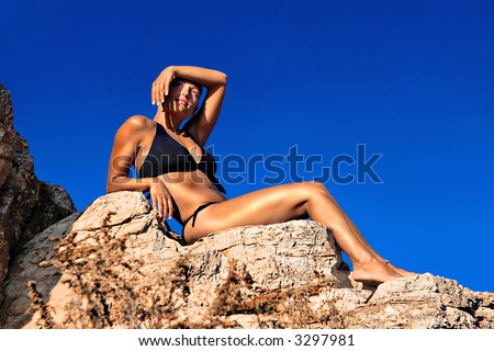 Young woman lying on rocks in black bikini shielding sun with hands
