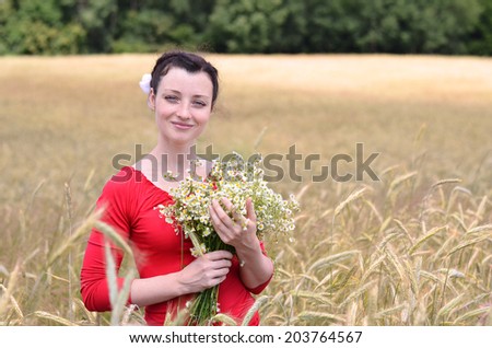 Attractive girl in red blouse standing in wheat field with bunch of flowers