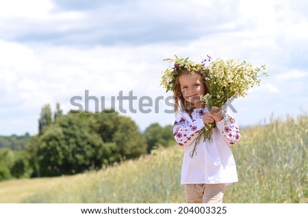 Beautiful little girl in Ukrainian traditional shirt standing in wheat field holding flowers