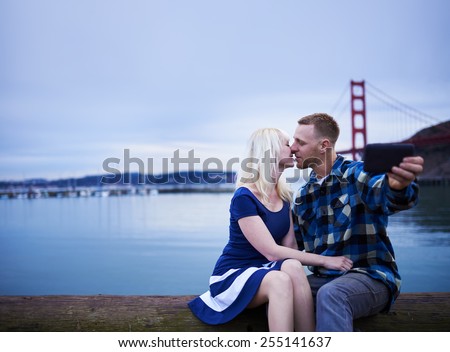 romantic couple kissing and taking selfie with smart phone at golden gate bridge with copy space and very cold/blue white balance to show day going to night
