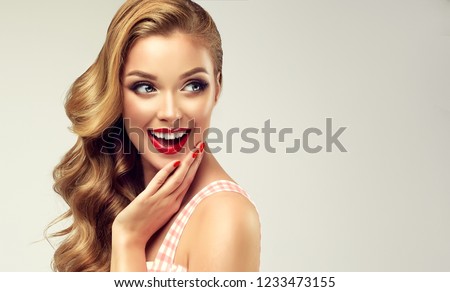 Woman with red lips and nails surprise holds cheeks by hand .Beautiful girl  with curly hair surprised and shocked   looking  aside . Presenting your product. Expressive facial expressions