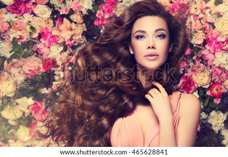 Brunette  girl with long  and   shiny wavy hair .  Beautiful  mode woman with curly hairstyle ,   background  wall of flowers .