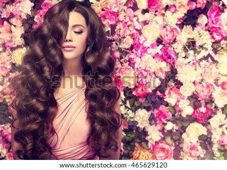 Brunette  girl with long  and   shiny wavy hair .  Beautiful  mode woman with curly hairstyle ,   background  wall of flowers .