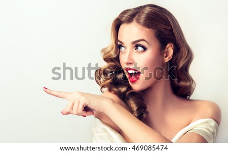 Woman surprise showing product .Beautiful girl  with curly hair  pointing to the side . Presenting your product. Isolated on white background. Expressive facial expressions