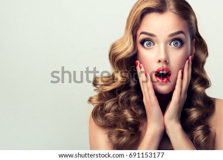 Woman with ren lips and nails surprise holds cheeks by hand .Beautiful girl  with curly hair surprised and shocked looks on you . Presenting your product. Expressive facial expressions