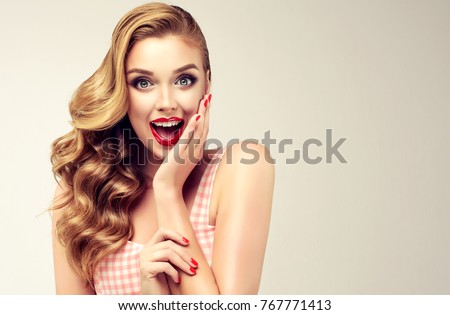 Woman with red lips and nails surprise holds cheeks by hand .Beautiful girl  with curly hair surprised and shocked looks on you . Presenting your product. Expressive facial expressions