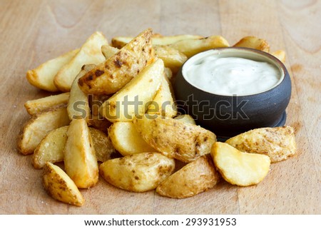 Heap of fried potato wedges on wood board with white dip in bowl.