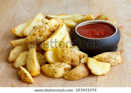 Heap of fried potato wedges on wood board with ketchup dip in bowl.