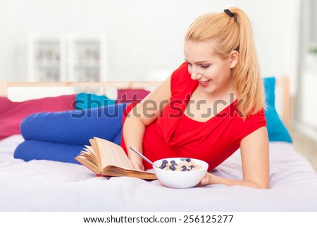 Young woman having breakfast and reading a book in bed.