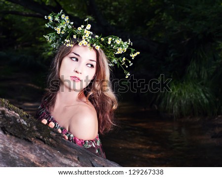 Beautiful girl in floral dress and wreath stand in the forest near river.