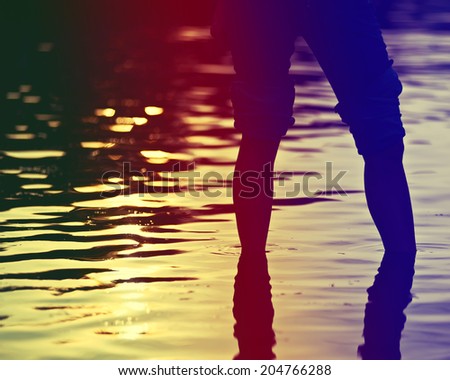 Female legs in rolled jeans in sea water on soft background of setting sun with retro effect filter.