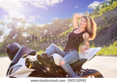 Young beautiful blonde girl in trendy jeans and a black t-shirt is resting on nature in seat of modern motorcycle. Outdoor portrait in soft sunny tones.