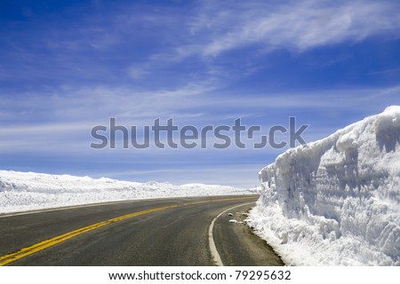 A highway that disappears around a corner is surrounded by snow walls that have been cut by a plow on a sunny day