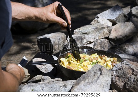 A hand holding a spatula, cooking breakfast over a campfire.