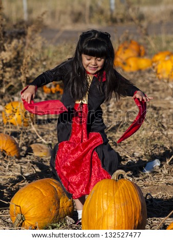 Little girl in Halloween costume looking for big pumpkin on pumpkin patch.