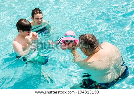 Family with cute baby girl faving fun in outdoor swimming pool on hot summer day.