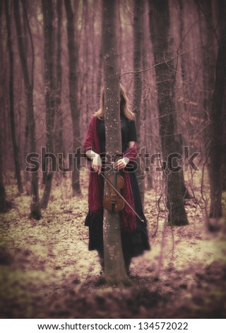 Fashion concept with woman in the forest behind a tree holding a violin and dressed in a black vintage dress