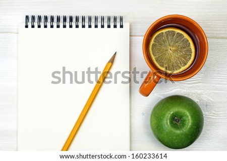 tea, green apple, pencil and notebook on a white wooden surface