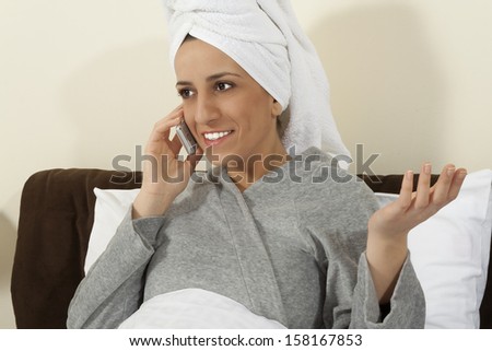 Young woman talking on mobile phone in bed with a towel on her head.