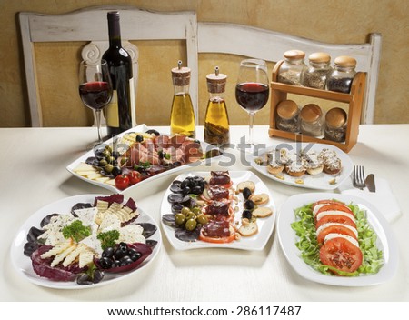 Five plates with appetizer, two glasses and a bottle of red wine, spice oil and jars with spices served on the table.