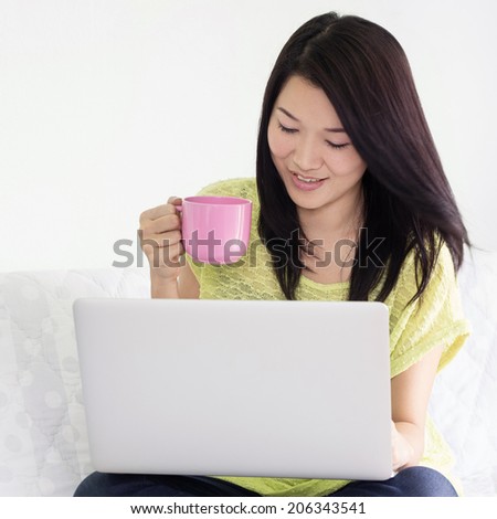 Happy young asian woman using laptop and holding pink cup of coffee indoor.