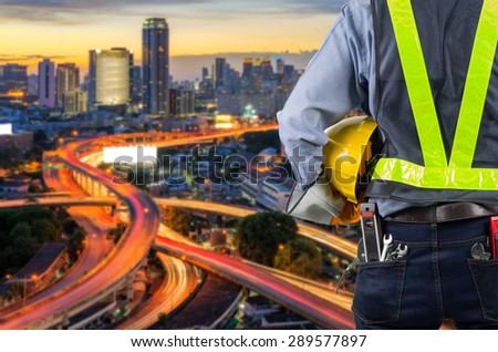 Engineer holding a yellow helmet for the safety of the workers, with blured light expressway as a backdrop and modern building in the business district twilight time.