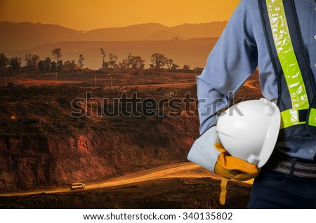 Engineer holding a white helmet for the safety of workers on a background of coal mining trucks are driving on the road. The sunset