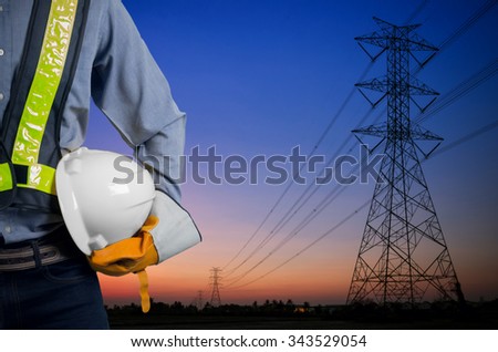 Engineer holding a white helmet for the safety of workers on the background. Silhouette transmission towers on the background of the evening sun.