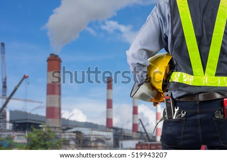 Engineer holding a yellow helmet with a coal power plant in the background.