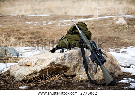 rifle with scope leaning up against a rock, backpack and box of ammunition