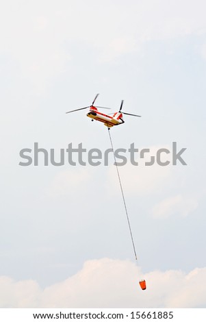 helicopter carrying water drop bucket, heavy twin-rotor aircraft fighting forest fire in US, motion blur on rotors