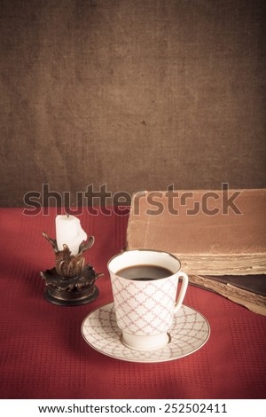 Stack of old books, cup of coffee, and ancient candle on the red tablecloth. Burlap background. Toned.