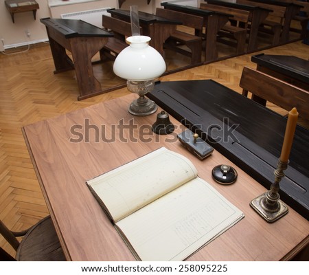 Classroom with parquet floor and rows of old desks. Table of teacher with journal, lamp, bell and candle.