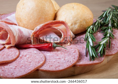 Slices of sausage and bacon on a cutting board with bread rolls, chilli pepper and rosemary.