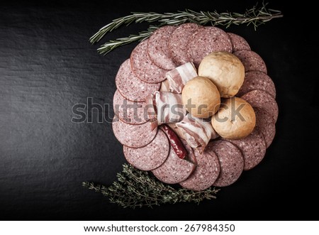 Slices of sausage and bacon on a cutting board with bread rolls, chilli pepper, thyme and rosemary on a black background. Toned.