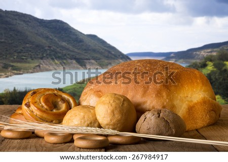 Bread assortment and wheat ears on an old wooden table opposite the mountains and river.
