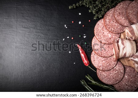 Slices of sausage and bacon on a round cutting board with rosemary, thyme, colored and chili pepper and sea salt on a black background. Toned.