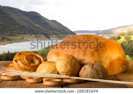 Bread assortment and wheat ears on an old wooden table opposite the mountains and river. Toned.