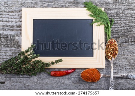 Herbs, chili, antique metal spoons with spices and black chalk board in a light wooden frame on the old wooden table.