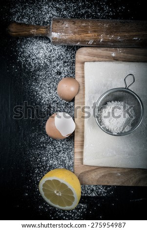 Set for home baking on a black background with flour. Rolling pin, dough, half of lemon, baking form, eggshell. Toned.