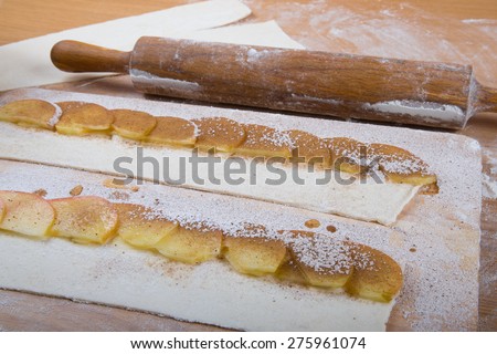 Apple slices on stripes of dough and rolling pin on a light wooden table with flour.