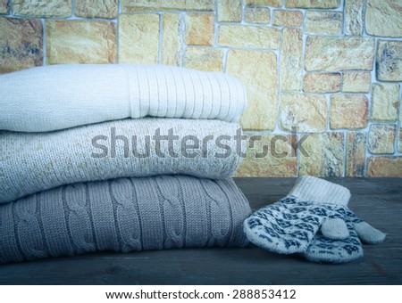 Stack of knitting clothes and painted mittens on wooden table opposite a stony wall. Toned.