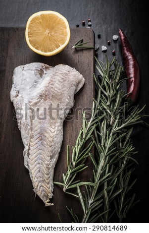 Fish filet, rosemary, red chili pepper, half a lemon and sea salt on brown cutting board on a black background. Toned.