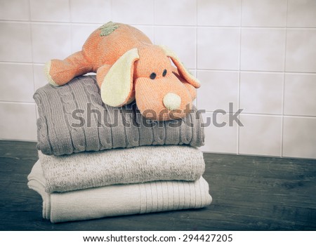 Knitted toy dog on the stack of knitted clothes on wooden table opposite a light tile wall. Toned.