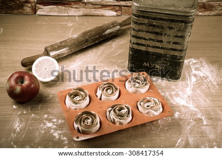 Silicone form with homemade pastries, a rolling pin, a jar with labeled FLOUR, half a lemon and an apple on a light wooden table. Toned