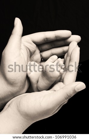 A young first time mother is holding the feet of her first new born baby girl and gently enclosing her soft and warm fingers and palms around the tiny feet and toes