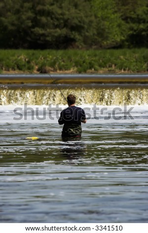 Man wading in a river, fly fishing with a low waterfall in the background.